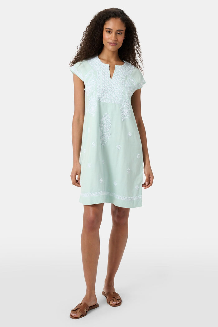 A woman with long curly hair wears the Roller Rabbit Faith Dress, a seaglass-green short-sleeved dress with hand embroidery and a notched neckline, standing against a plain light background.