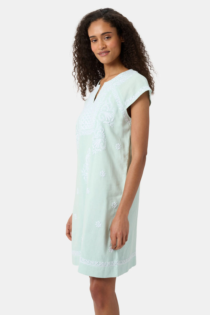 A woman with long curly hair wears the Roller Rabbit Faith Dress, a seaglass-green short-sleeved dress with hand embroidery and a notched neckline, standing against a plain light background.