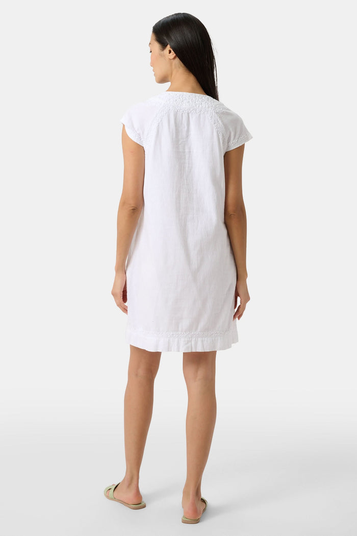 A woman stands against a plain light background, wearing the Roller Rabbit Faith Dress—a short-sleeve, knee-length white embroidered dress with a keyhole neckline—paired with green sandals.