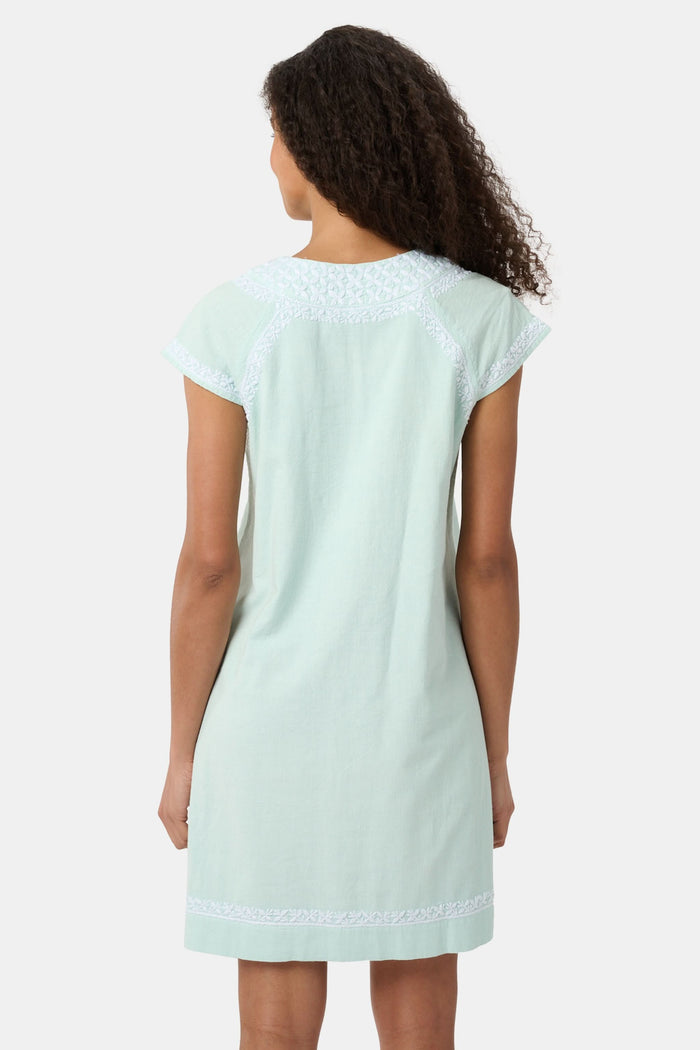 A woman with long curly hair wears the Roller Rabbit Faith Dress, a seaglass-green short-sleeved dress with hand embroidery and a notched neckline, standing against a plain light background.