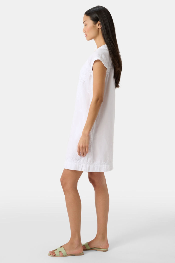 A woman stands against a plain light background, wearing the Roller Rabbit Faith Dress—a short-sleeve, knee-length white embroidered dress with a keyhole neckline—paired with green sandals.