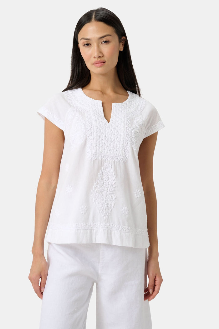 A woman with long dark hair wears the Roller Rabbit Faith Top, a white embroidered cotton blouse with short sleeves and a keyhole neckline, paired with white pants, standing against a plain light background.