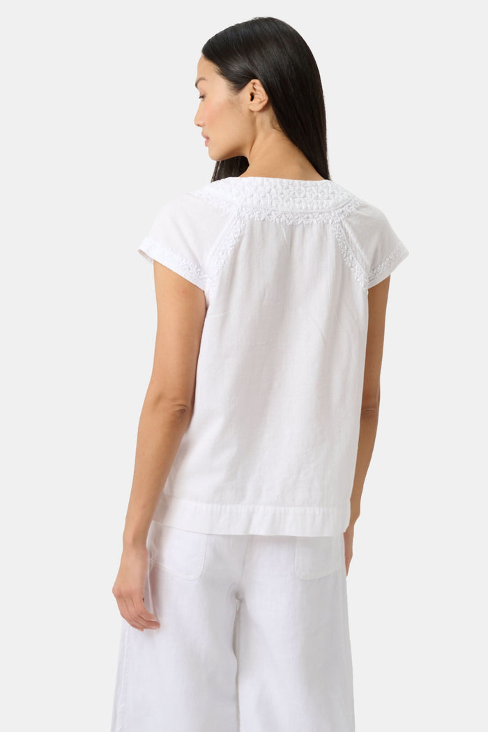 A woman with long dark hair wears the Roller Rabbit Faith Top, a white embroidered cotton blouse with short sleeves and a keyhole neckline, paired with white pants, standing against a plain light background.