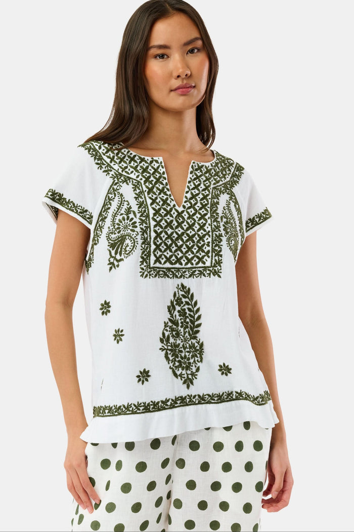 A woman wearing sunglasses, the Roller Rabbit Faith Top—a hand-embroidered white cotton blouse with green details—and a matching polka dot scarf sits by the water on a sunny day.
