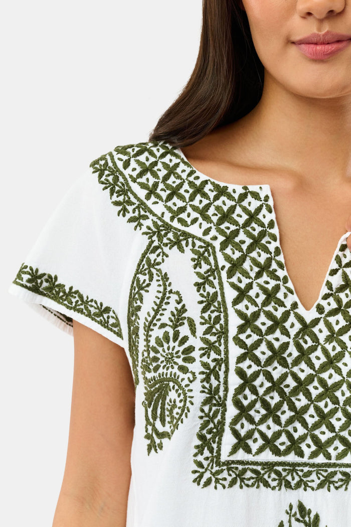 A woman wearing sunglasses, the Roller Rabbit Faith Top—a hand-embroidered white cotton blouse with green details—and a matching polka dot scarf sits by the water on a sunny day.