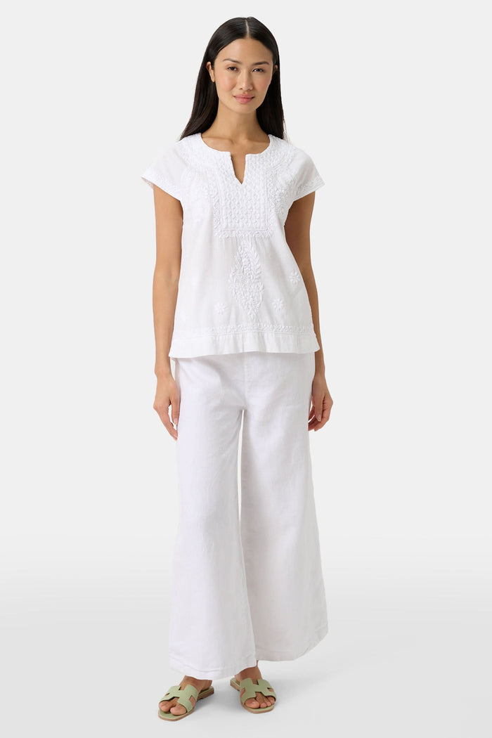 A woman with long dark hair wears the Roller Rabbit Faith Top, a white embroidered cotton blouse with short sleeves and a keyhole neckline, paired with white pants, standing against a plain light background.