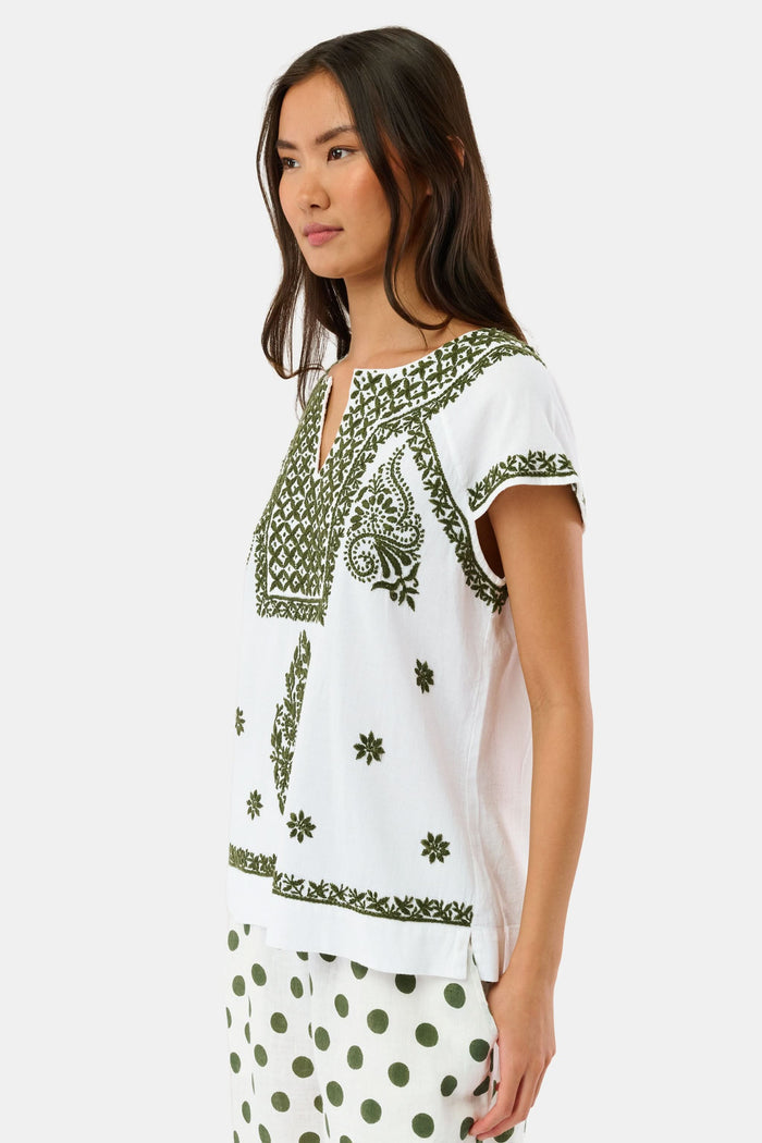 A woman wearing sunglasses, the Roller Rabbit Faith Top—a hand-embroidered white cotton blouse with green details—and a matching polka dot scarf sits by the water on a sunny day.