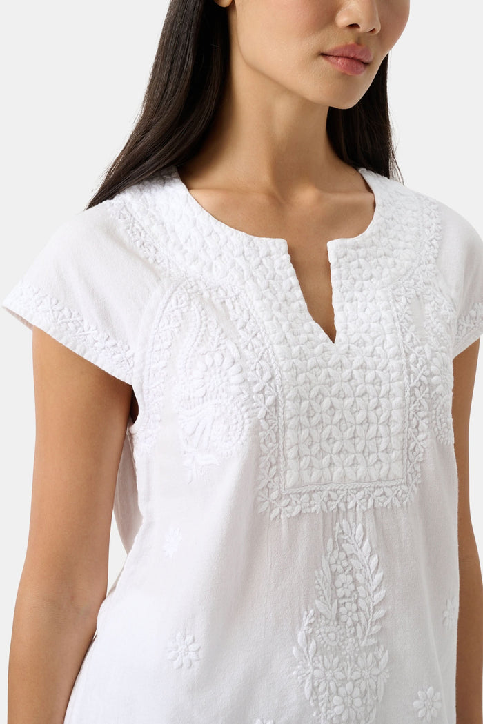 A woman with long dark hair wears the Roller Rabbit Faith Top, a white embroidered cotton blouse with short sleeves and a keyhole neckline, paired with white pants, standing against a plain light background.