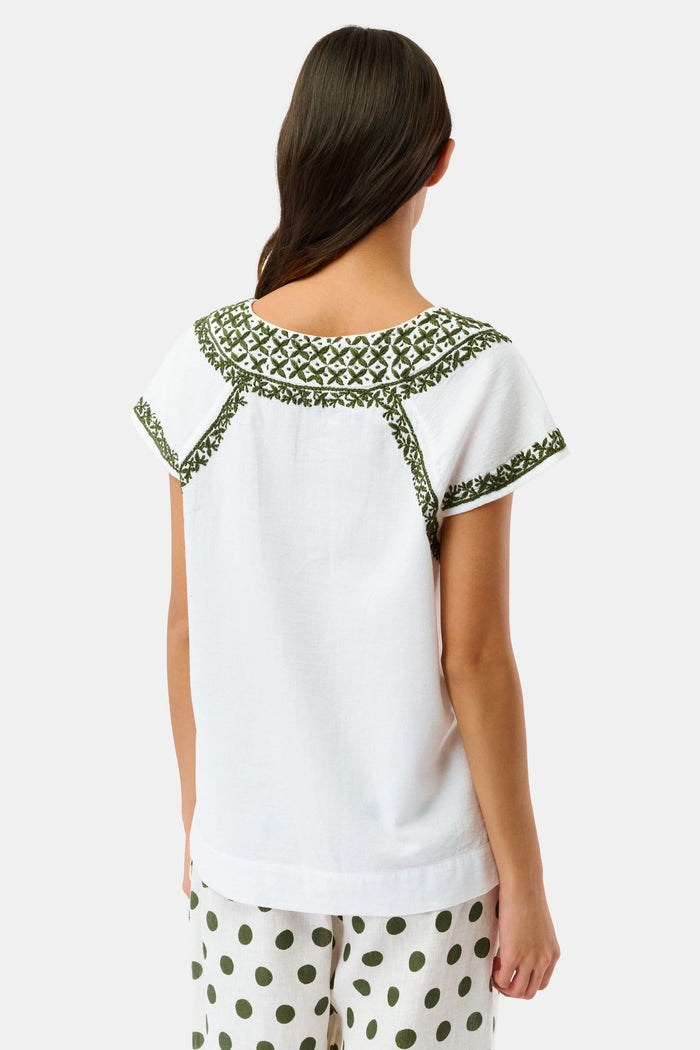 A woman wearing sunglasses, the Roller Rabbit Faith Top—a hand-embroidered white cotton blouse with green details—and a matching polka dot scarf sits by the water on a sunny day.