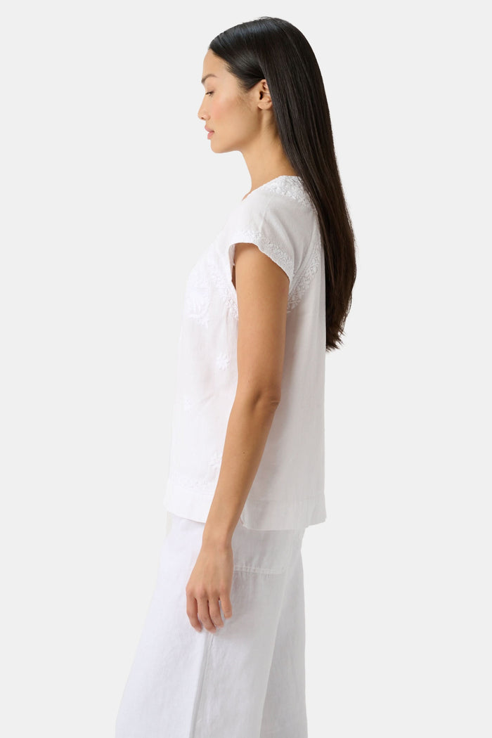 A woman with long dark hair wears the Roller Rabbit Faith Top, a white embroidered cotton blouse with short sleeves and a keyhole neckline, paired with white pants, standing against a plain light background.