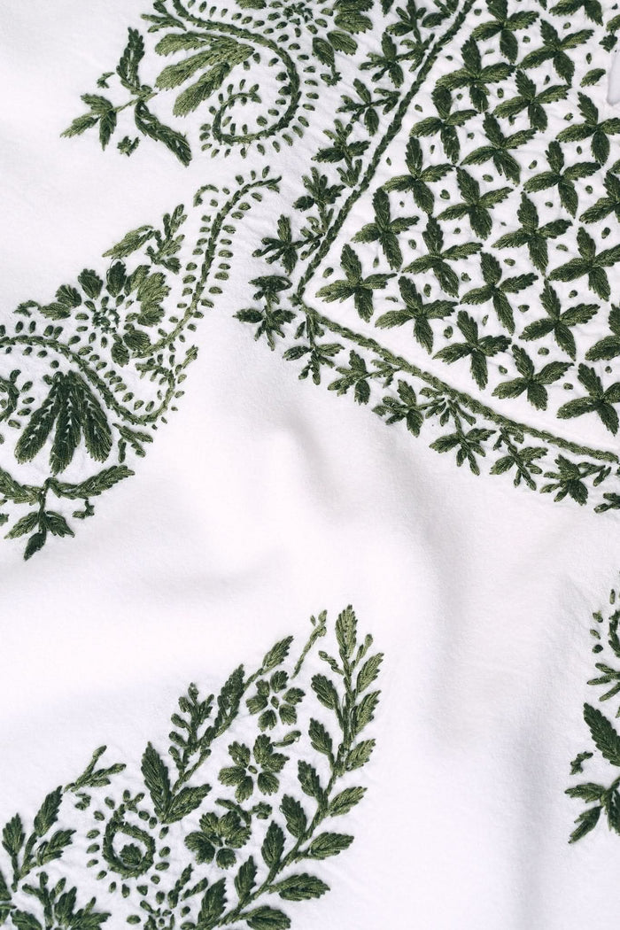 A woman wearing sunglasses, the Roller Rabbit Faith Top—a hand-embroidered white cotton blouse with green details—and a matching polka dot scarf sits by the water on a sunny day.
