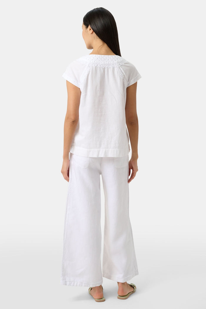 A woman with long dark hair wears the Roller Rabbit Faith Top, a white embroidered cotton blouse with short sleeves and a keyhole neckline, paired with white pants, standing against a plain light background.
