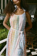 A woman stands on a checkered floor, wearing the Roller Rabbit Fan Geo Courtney Dress—a multicolored cotton voile maxi with a white rope belt and matching headband. Her curly hair is partly covered, and striped canopies are visible in the background.