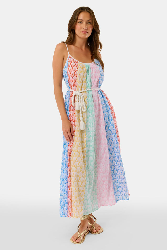 A woman stands on a checkered floor, wearing the Roller Rabbit Fan Geo Courtney Dress—a multicolored cotton voile maxi with a white rope belt and matching headband. Her curly hair is partly covered, and striped canopies are visible in the background.