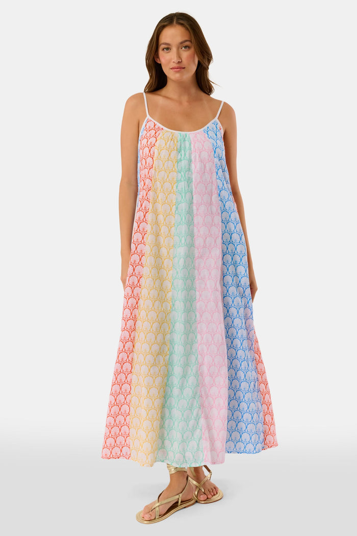 A woman stands on a checkered floor, wearing the Roller Rabbit Fan Geo Courtney Dress—a multicolored cotton voile maxi with a white rope belt and matching headband. Her curly hair is partly covered, and striped canopies are visible in the background.
