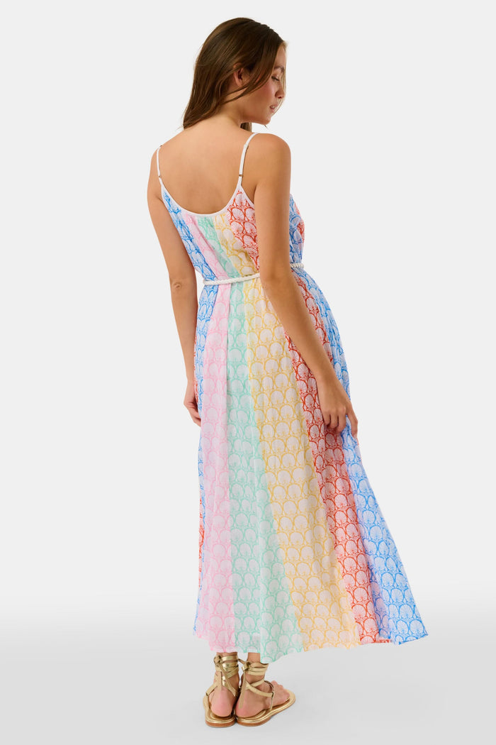 A woman stands on a checkered floor, wearing the Roller Rabbit Fan Geo Courtney Dress—a multicolored cotton voile maxi with a white rope belt and matching headband. Her curly hair is partly covered, and striped canopies are visible in the background.