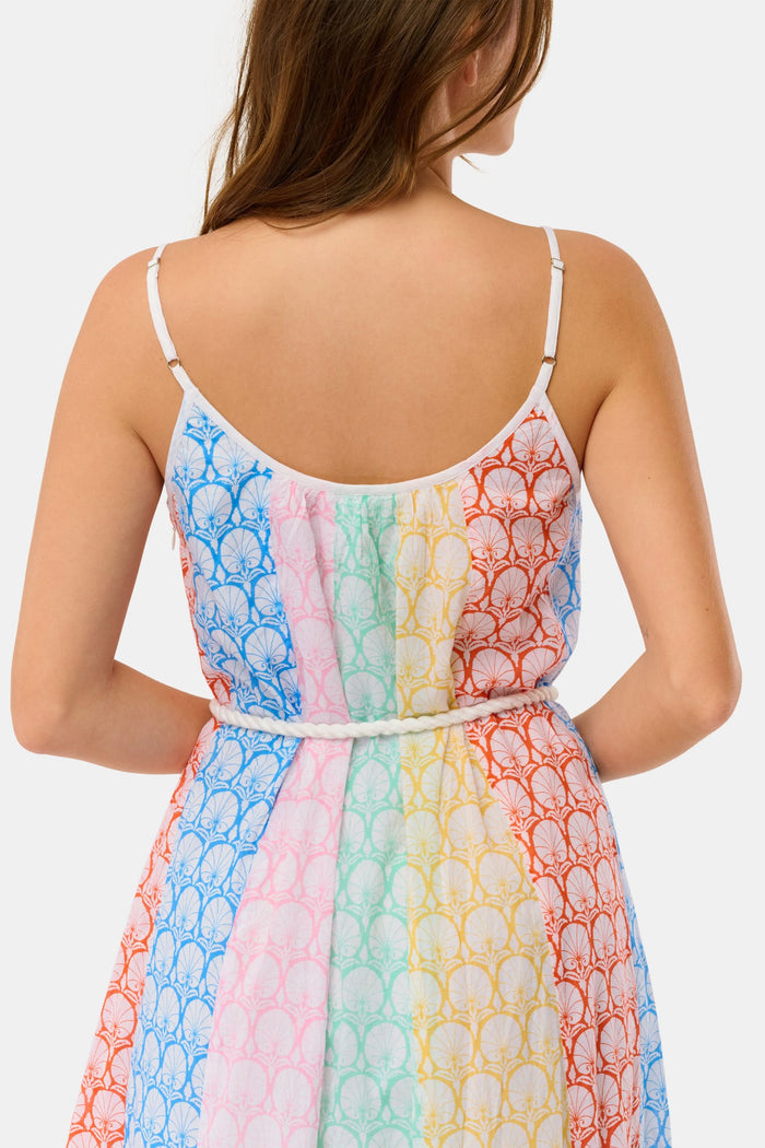 A woman stands on a checkered floor, wearing the Roller Rabbit Fan Geo Courtney Dress—a multicolored cotton voile maxi with a white rope belt and matching headband. Her curly hair is partly covered, and striped canopies are visible in the background.