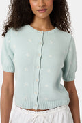 Close-up of a woman wearing the Roller Rabbit Floral Bloom Nyla Cardigan, a light blue cotton button-up with white embroidered stars, rounded neckline, and short sleeves, styled with white eyelet shorts.