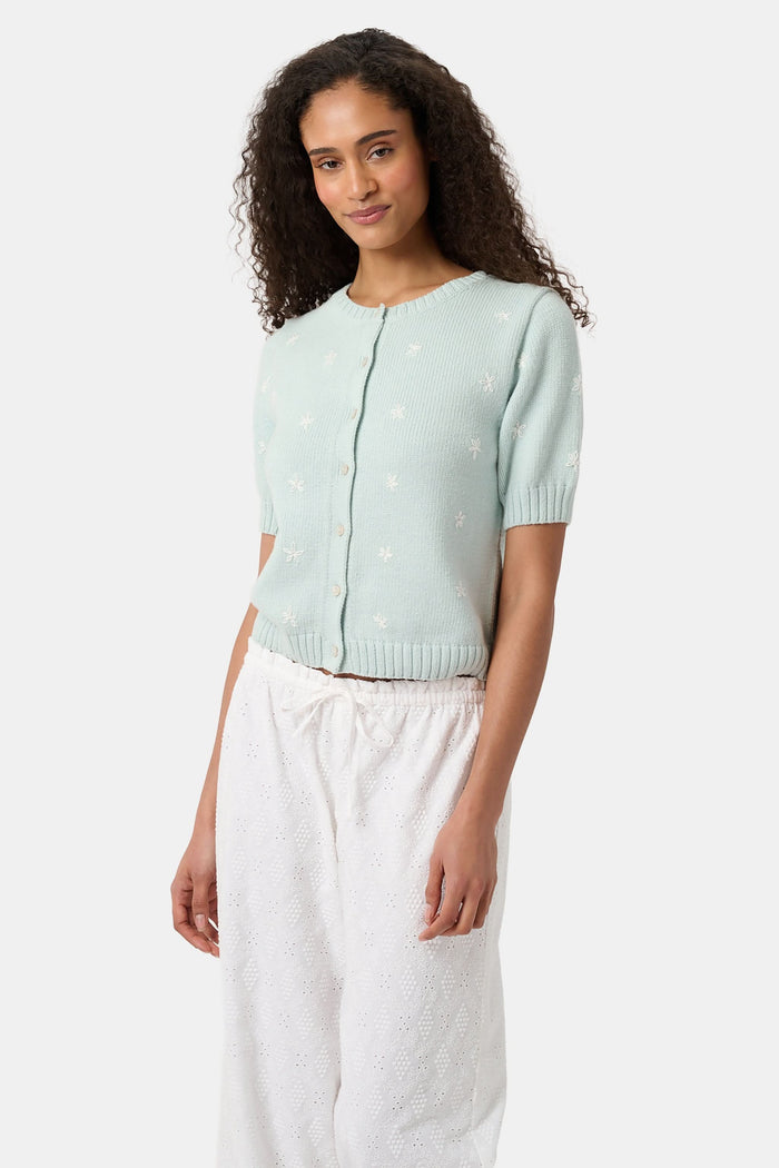 Close-up of a woman wearing the Roller Rabbit Floral Bloom Nyla Cardigan, a light blue cotton button-up with white embroidered stars, rounded neckline, and short sleeves, styled with white eyelet shorts.