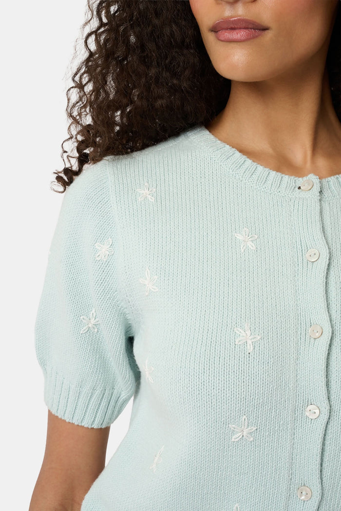 Close-up of a woman wearing the Roller Rabbit Floral Bloom Nyla Cardigan, a light blue cotton button-up with white embroidered stars, rounded neckline, and short sleeves, styled with white eyelet shorts.