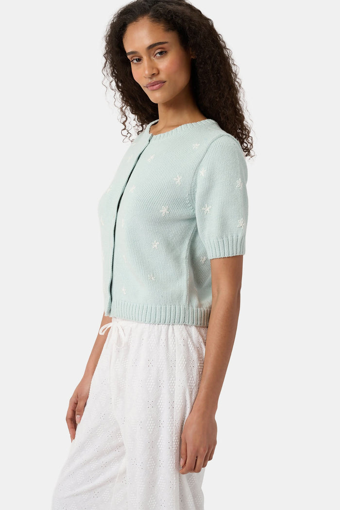 Close-up of a woman wearing the Roller Rabbit Floral Bloom Nyla Cardigan, a light blue cotton button-up with white embroidered stars, rounded neckline, and short sleeves, styled with white eyelet shorts.