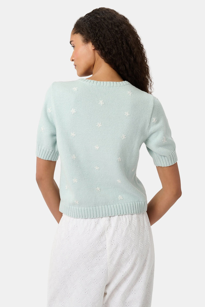 Close-up of a woman wearing the Roller Rabbit Floral Bloom Nyla Cardigan, a light blue cotton button-up with white embroidered stars, rounded neckline, and short sleeves, styled with white eyelet shorts.