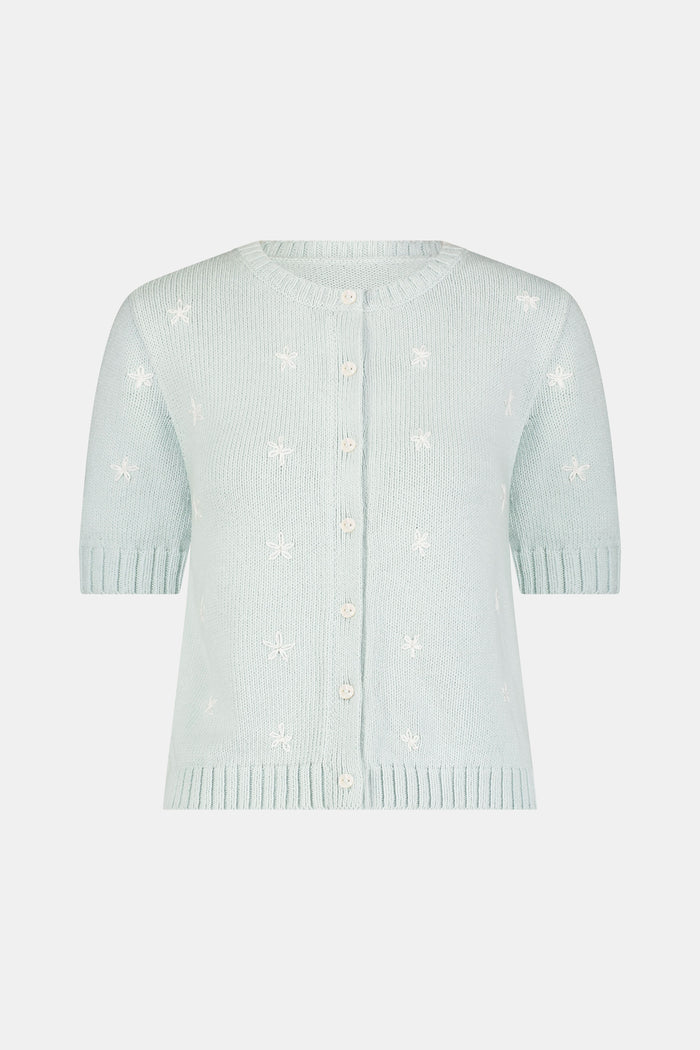 Close-up of a woman wearing the Roller Rabbit Floral Bloom Nyla Cardigan, a light blue cotton button-up with white embroidered stars, rounded neckline, and short sleeves, styled with white eyelet shorts.