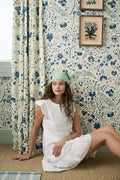 A woman in a white Florence Lace Pippa Dress by Roller Rabbit and a green headscarf sits on the floor against blue floral wallpaper, near a window with matching curtains and framed botanical art.