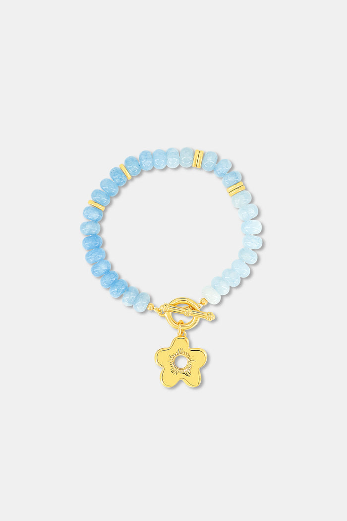 The Roller Rabbit Flower Charm Beaded Bracelet features light blue round beads with gold spacers, a gold flower-shaped charm, and a gold toggle clasp, displayed on a plain white background.