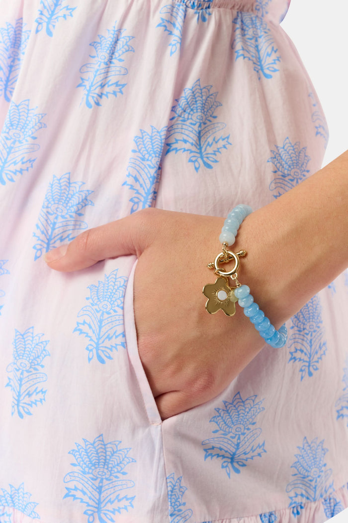The Roller Rabbit Flower Charm Beaded Bracelet features light blue round beads with gold spacers, a gold flower-shaped charm, and a gold toggle clasp, displayed on a plain white background.