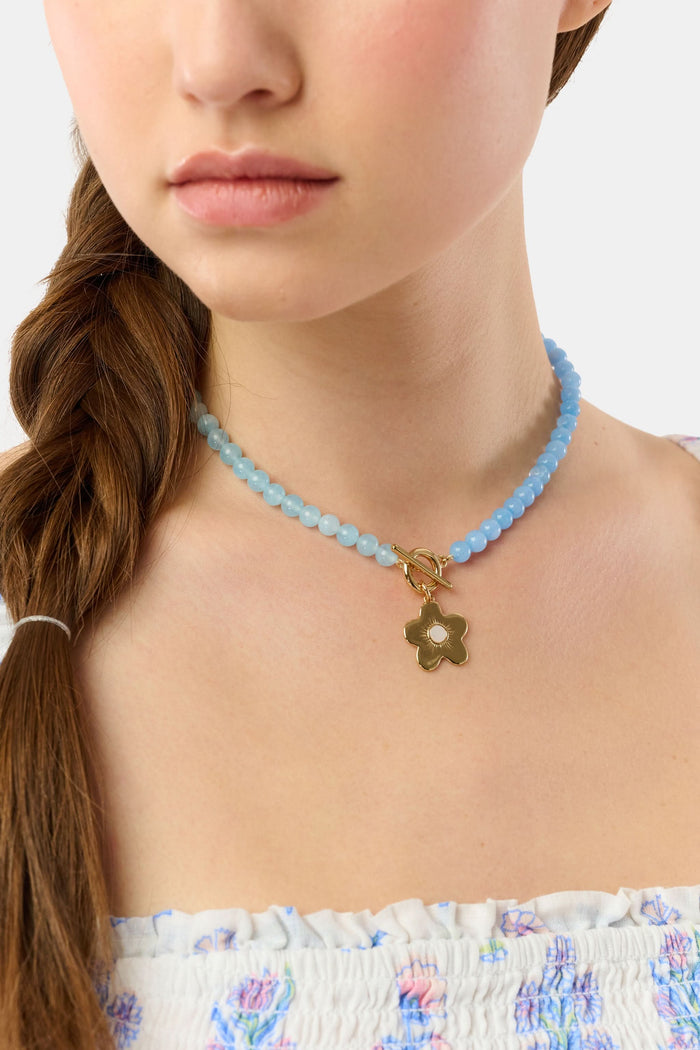 A woman with a brown braid models Roller Rabbit's Flower Charm Beaded Necklace, a blue beaded piece with a gold flower charm. She wears a white floral smocked top; only her lower face, neck, and shoulders are shown.