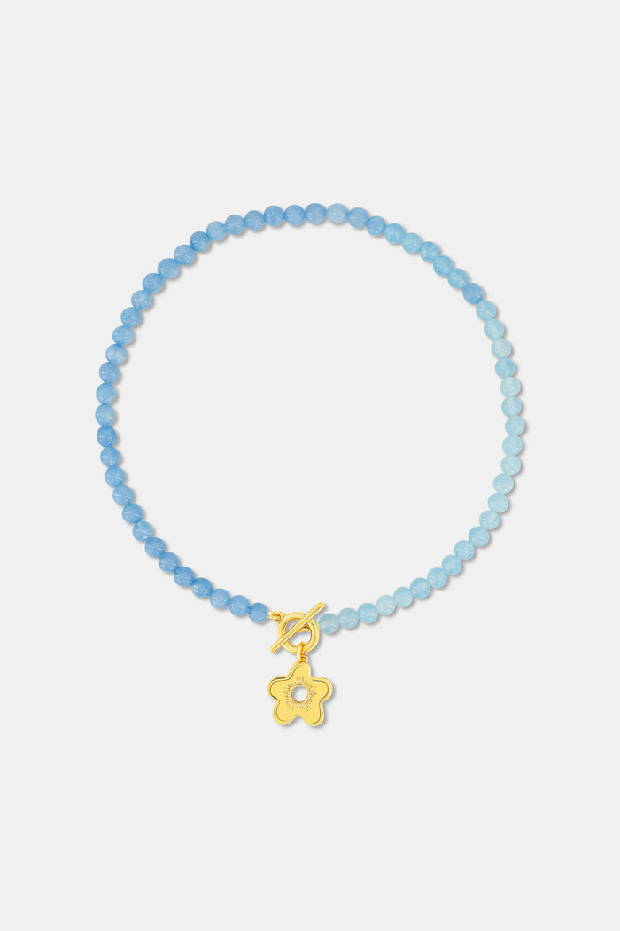 A woman with a brown braid models Roller Rabbit's Flower Charm Beaded Necklace, a blue beaded piece with a gold flower charm. She wears a white floral smocked top; only her lower face, neck, and shoulders are shown.