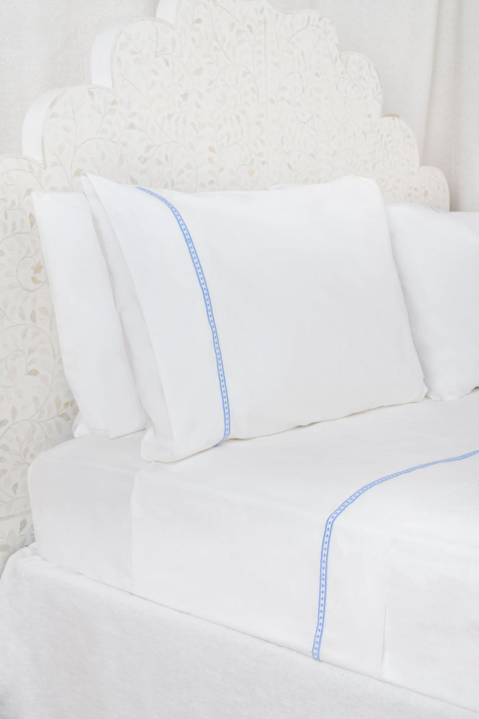 A neatly made bed features the Roller Rabbit Francis Embroidered Sheet Set—white cotton sheets and pillowcases with thin blue embroidery. The headboard is upholstered with a white, floral-patterned fabric.