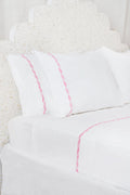 The Francis Embroidered Sheet Set by Roller Rabbit features white cotton sheets and pillowcases with a thin pink border, perfectly complementing an ornate white patterned headboard for a clean, elegant look.