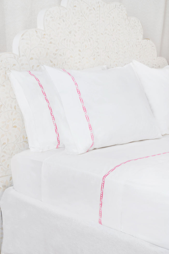 The Francis Embroidered Sheet Set by Roller Rabbit features white cotton sheets and pillowcases with a thin pink border, perfectly complementing an ornate white patterned headboard for a clean, elegant look.