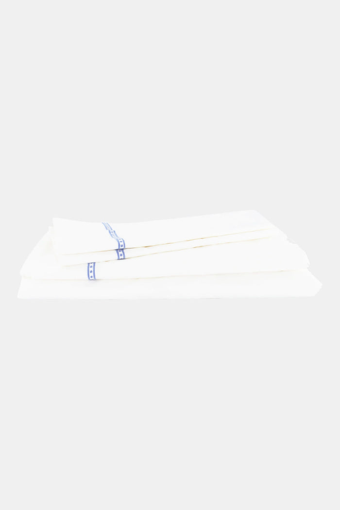 A neatly made bed features the Roller Rabbit Francis Embroidered Sheet Set—white cotton sheets and pillowcases with thin blue embroidery. The headboard is upholstered with a white, floral-patterned fabric.