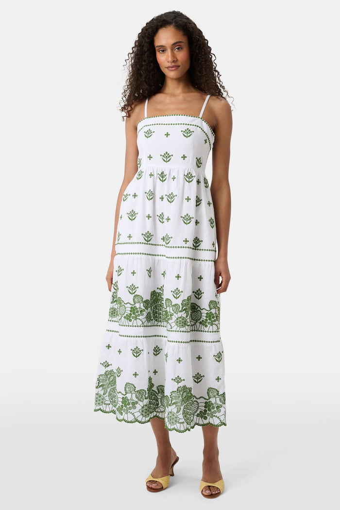 A woman with curly hair wears the Roller Rabbit Fruit Eyelet Selena Dress, a green linen maxi with floral embroidery and thin straps. She stands facing forward in yellow open-toe sandals against a plain white background.