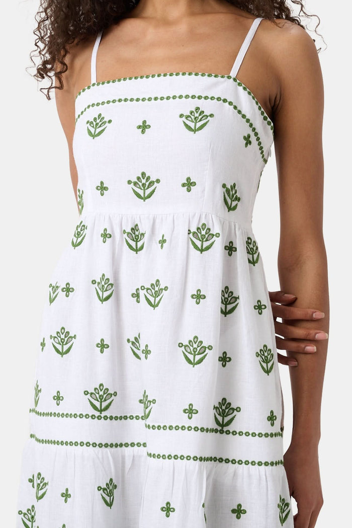 A woman with curly hair wears the Roller Rabbit Fruit Eyelet Selena Dress, a green linen maxi with floral embroidery and thin straps. She stands facing forward in yellow open-toe sandals against a plain white background.