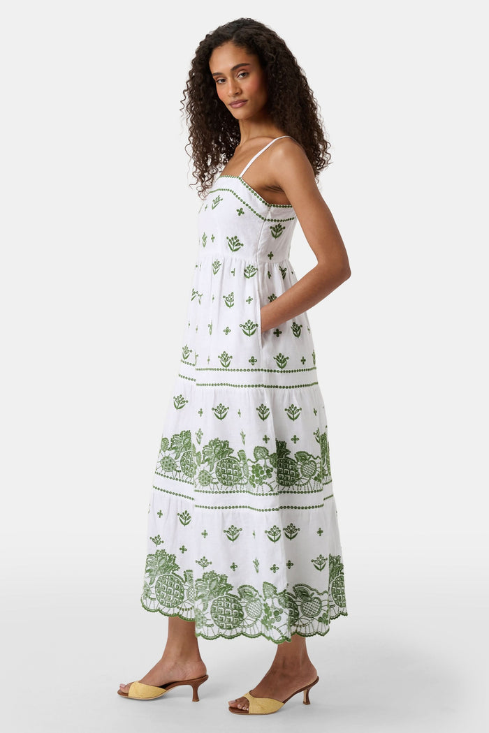 A woman with curly hair wears the Roller Rabbit Fruit Eyelet Selena Dress, a green linen maxi with floral embroidery and thin straps. She stands facing forward in yellow open-toe sandals against a plain white background.