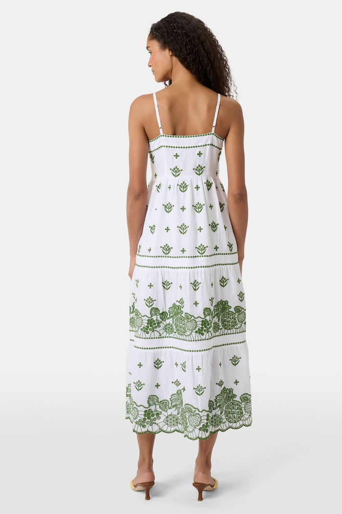 A woman with curly hair wears the Roller Rabbit Fruit Eyelet Selena Dress, a green linen maxi with floral embroidery and thin straps. She stands facing forward in yellow open-toe sandals against a plain white background.