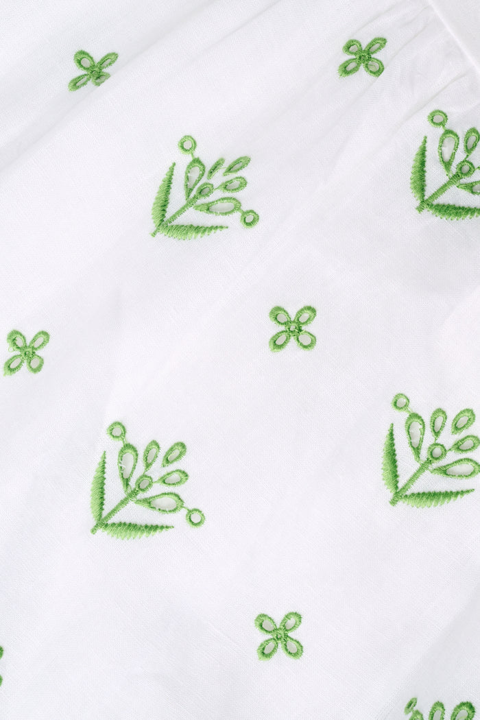 A woman with curly hair wears the Roller Rabbit Fruit Eyelet Selena Dress, a green linen maxi with floral embroidery and thin straps. She stands facing forward in yellow open-toe sandals against a plain white background.