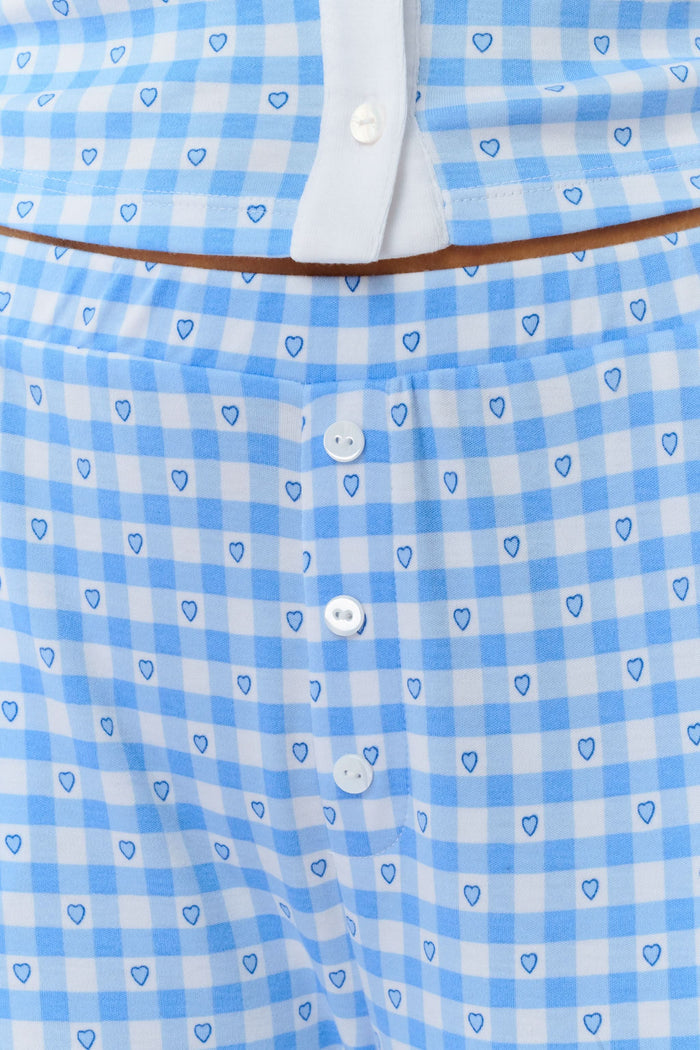 A person stands barefoot wearing Roller Rabbit’s Gingham Hearts Boden Pant—light blue, wide-leg Pima cotton pants with an elastic waist—and a matching long-sleeve top with blue gingham hearts. Their hands rest on their hips. Head and feet are out of frame.