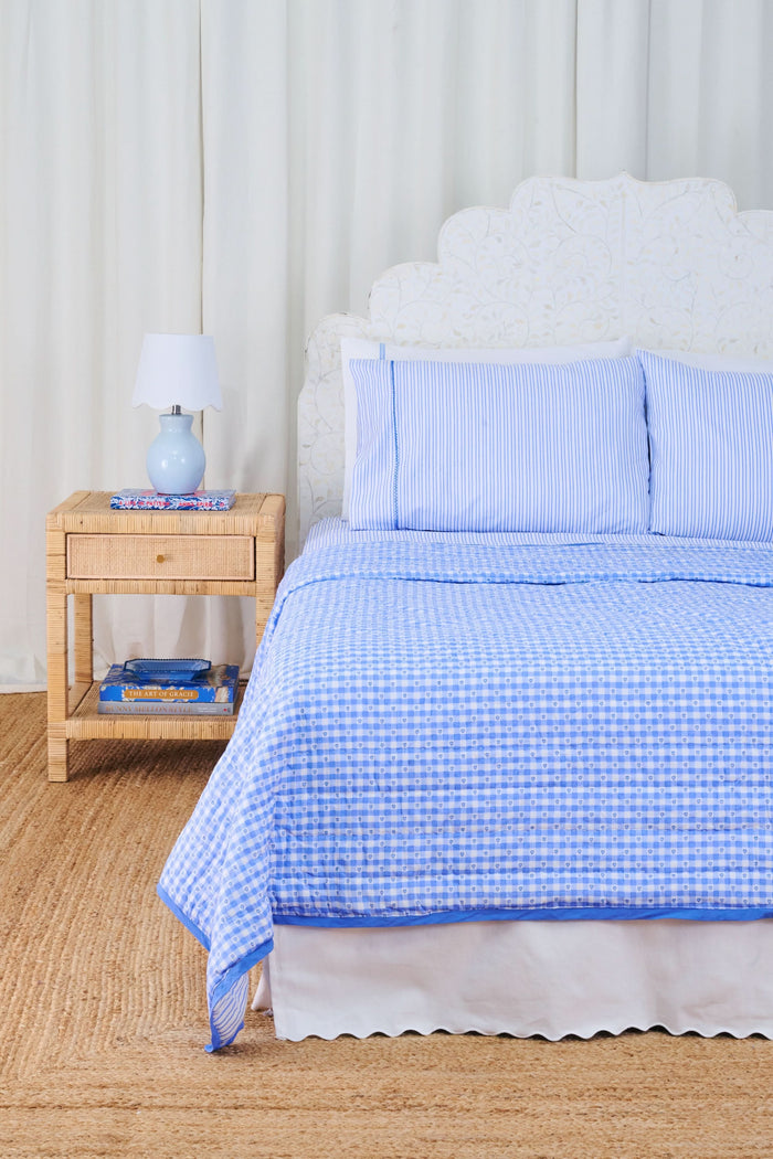 A bed dressed in the Roller Rabbit Gingham Hearts/Capellini Quilt with gingham heart pillows sits beside a wicker nightstand, blue lamp, books, and light curtains, all atop a woven rug.