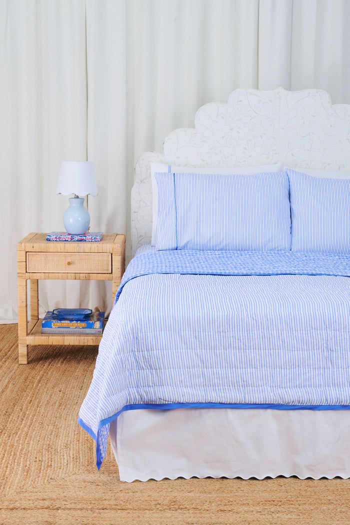 A bed dressed in the Roller Rabbit Gingham Hearts/Capellini Quilt with gingham heart pillows sits beside a wicker nightstand, blue lamp, books, and light curtains, all atop a woven rug.