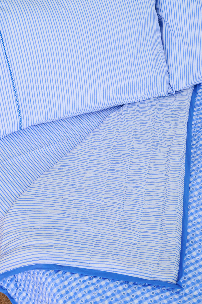 A bed dressed in the Roller Rabbit Gingham Hearts/Capellini Quilt with gingham heart pillows sits beside a wicker nightstand, blue lamp, books, and light curtains, all atop a woven rug.