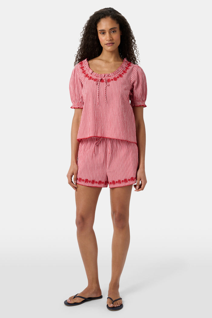 A person is wearing the Roller Rabbit Gingham James Short—red and white cotton gingham with an elastic drawstring waist, pockets, and red embroidered floral trim—paired with a matching short-sleeve top. Their left hand is in their pocket.