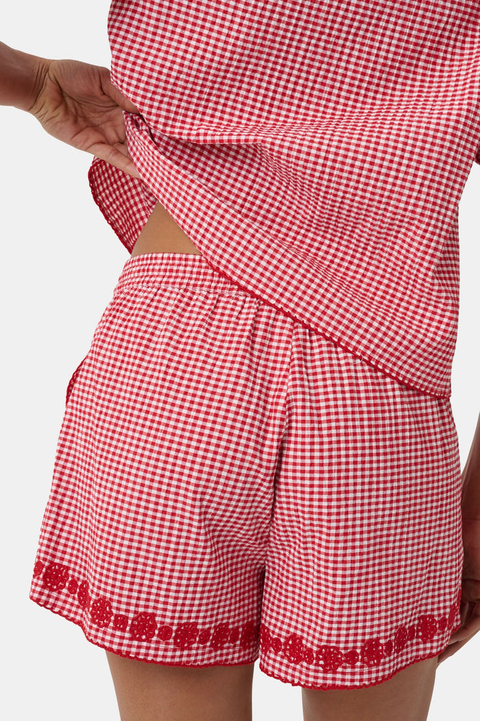 A person is wearing the Roller Rabbit Gingham James Short—red and white cotton gingham with an elastic drawstring waist, pockets, and red embroidered floral trim—paired with a matching short-sleeve top. Their left hand is in their pocket.