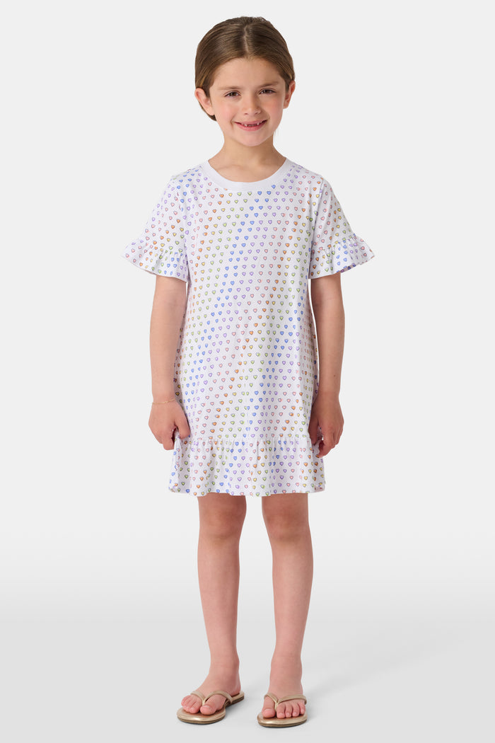 A young child smiles in the Roller Rabbit Girls Disco Hearts Esme Dress, a short-sleeve cotton dress with a ruffled hem and colorful heart patterns, set against a plain, light background.