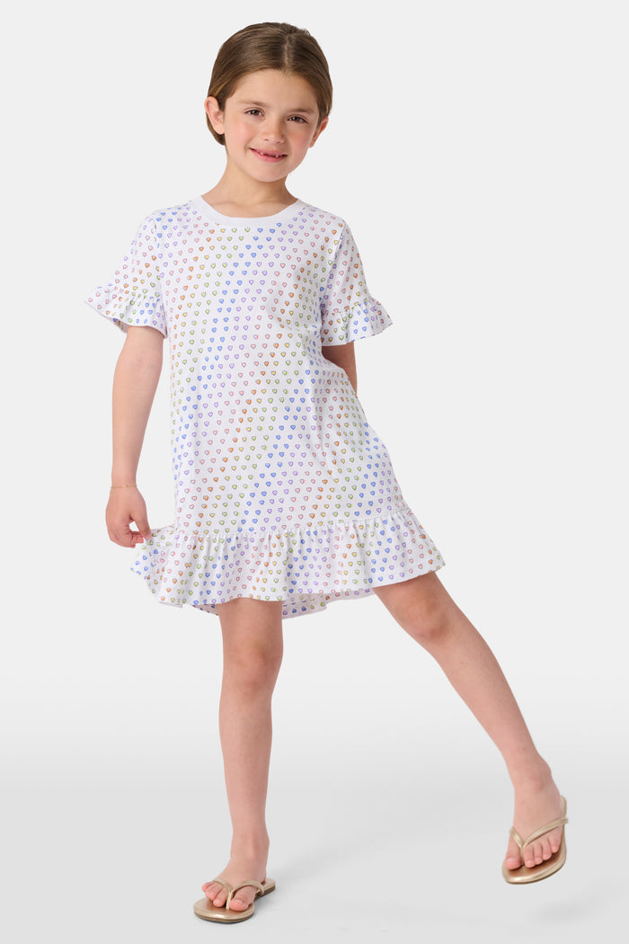 A young child smiles in the Roller Rabbit Girls Disco Hearts Esme Dress, a short-sleeve cotton dress with a ruffled hem and colorful heart patterns, set against a plain, light background.