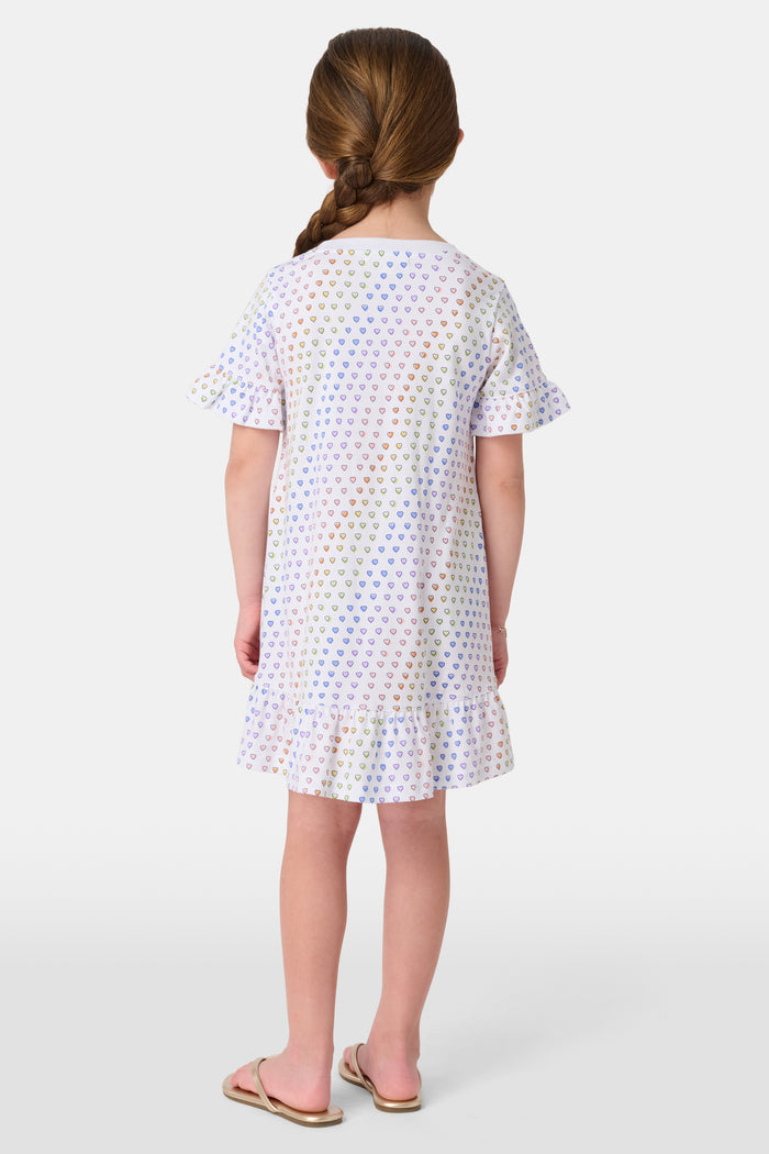 A young child smiles in the Roller Rabbit Girls Disco Hearts Esme Dress, a short-sleeve cotton dress with a ruffled hem and colorful heart patterns, set against a plain, light background.